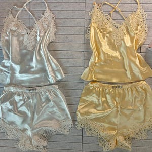 May include: Two sets of satin lingerie. One set is ivory, the other is yellow. Each set includes a camisole with lace trim and shorts with lace edging. The camisoles have a criss-cross back design. A gift box with a card is visible.