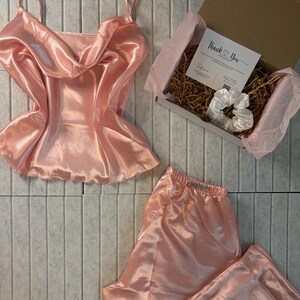 Op de afbeelding: Een lichtroze satijnen pyjamaset, bestaande uit een camisole top en een broek, wordt getoond. Een geschenkdoos met een scrunchie en een bedankkaart is ook zichtbaar. De set is gemaakt van een glanzende, gladde stof.