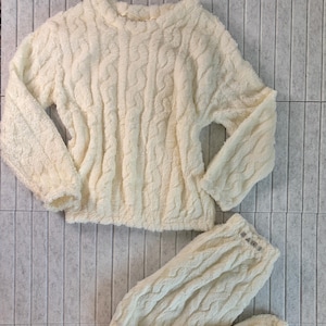 May include: Cream-colored, cable-knit pajama set. The set includes a long-sleeved top and matching pants. The fabric appears soft and plush, ideal for lounging. The top has a crew neck, and the pants have a drawstring waist.