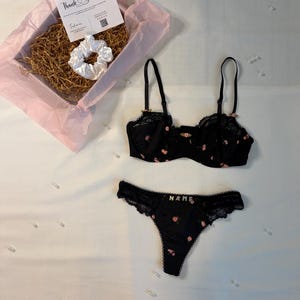 May include: A black lingerie set with pink floral accents, including a bra and thong. The thong has the word "NAME" embroidered on it. A white scrunchie and a gift box with a "Thank You" card are also present.