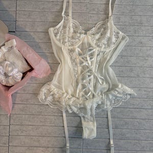 May include: An ivory-coloured lingerie set. The bodysuit has a lace-trimmed design, corset-style front, and adjustable straps. The set includes a matching garter belt. The items are displayed on a grey surface.