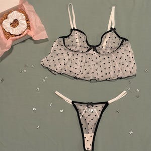 May include: A white lingerie set with black polka dots, featuring a bralette and thong. The thong has the word "NAME" printed on it. A white scrunchie is in a box with pink tissue paper. The set is on a green surface.