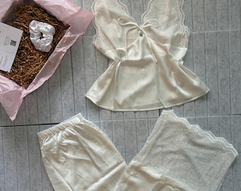 May include: A cream-colored two-piece pajama set consisting of a sleeveless camisole with a lace V-neckline and a tie-front detail, paired with matching wide-leg trousers featuring lace trim at the hem. The set includes a white satin hair scrunchie.