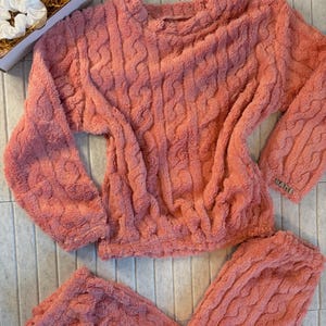 May include: A coral-colored, cable-knit pajama set. The set includes a long-sleeved top and matching pants. The top has a crew neck and the word "WARE" is embroidered on the sleeve. A white scrunchie and a box are in the background.