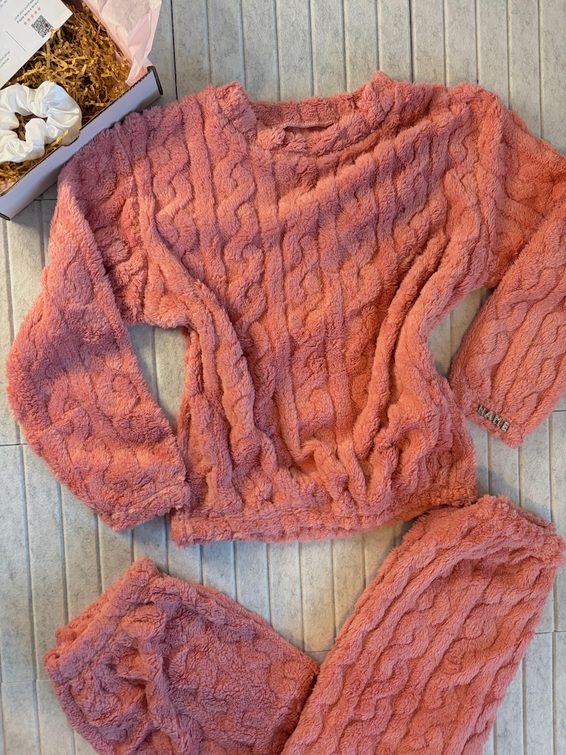 May include: A coral-colored, cable-knit pajama set. The set includes a long-sleeved top and matching pants. The top has a crew neck and the word "WARE" is embroidered on the sleeve. A white scrunchie and a box are in the background.