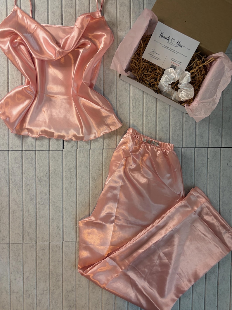 Op de afbeelding: Een lichtroze satijnen pyjamaset, bestaande uit een camisole top en een broek, wordt getoond. Een geschenkdoos met een scrunchie en een bedankkaart is ook zichtbaar. De set is gemaakt van een glanzende, gladde stof.