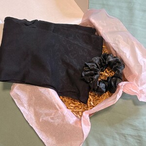 May include: A black pair of shorts and a black scrunchie are nestled inside a cardboard box lined with pink tissue paper and gold-colored paper filler. The box is open, revealing the contents.