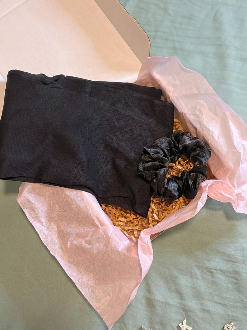 May include: A black pair of shorts and a black scrunchie are nestled inside a cardboard box lined with pink tissue paper and gold-colored paper filler. The box is open, revealing the contents.
