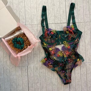 May include: A dark green bodysuit with floral embroidery in yellow, red, and purple. The lingerie has adjustable straps and a scalloped edge. A box with a scrunchie is also visible.