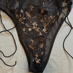 May include: Black lingerie with gold floral embroidery. The sheer fabric is adorned with detailed, embroidered flowers and vines. The garment features a high-cut leg and delicate tie straps for an adjustable fit. A small, silver-colored charm is visible.