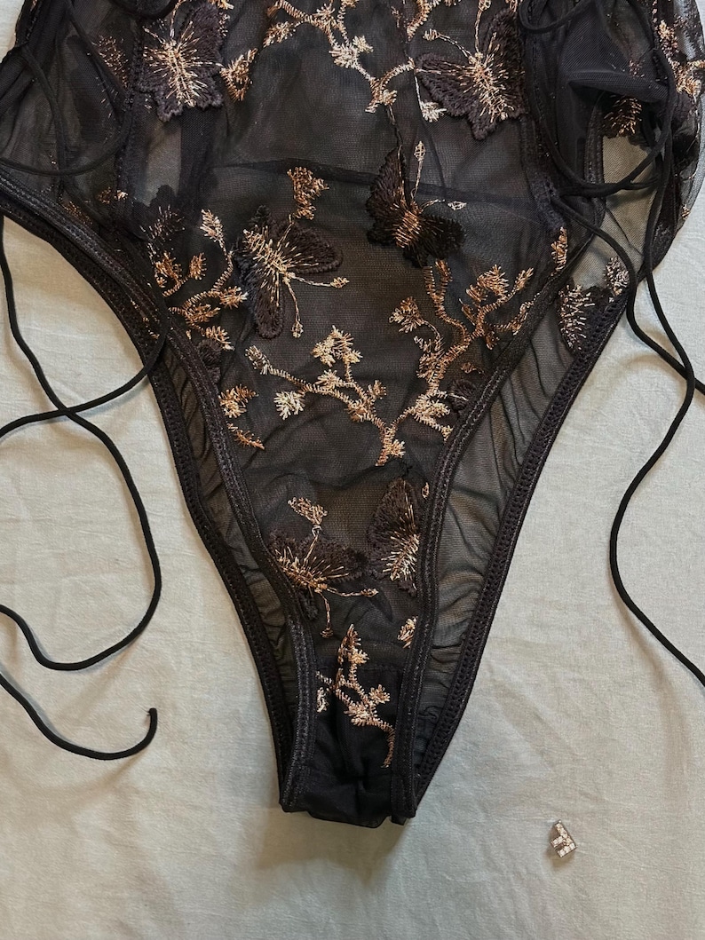 May include: Black lingerie with gold floral embroidery. The sheer fabric is adorned with detailed, embroidered flowers and vines. The garment features a high-cut leg and delicate tie straps for an adjustable fit. A small, silver-colored charm is visible.