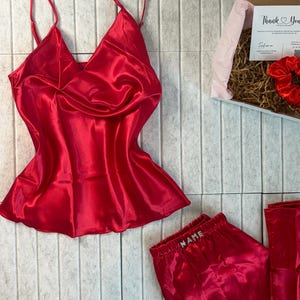 May include: A red satin pajama set, including a camisole top with spaghetti straps and a pair of pajama pants. The pants have the word "NAME" embroidered on the waistband. A gift box with a card and a scrunchie is also visible.