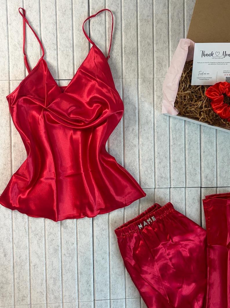 May include: A red satin pajama set, including a camisole top with spaghetti straps and a pair of pajama pants. The pants have the word "NAME" embroidered on the waistband. A gift box with a card and a scrunchie is also visible.
