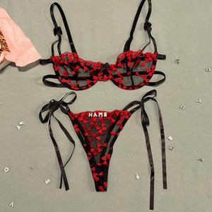 Op de afbeelding: Een zwarte lingerieset bestaande uit een transparante mesh basis met rode hartvormige borduursels. De bh heeft verstelbare bandjes en beugels. De bijpassende string heeft zijstrikjes en het woord NAME in zilverkleurige blokletters.
