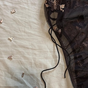 May include: A sheer black lingerie piece with gold butterfly and floral embroidery. The garment features delicate lace detailing and a drawstring. Scattered around are small silver-colored decorative charms.