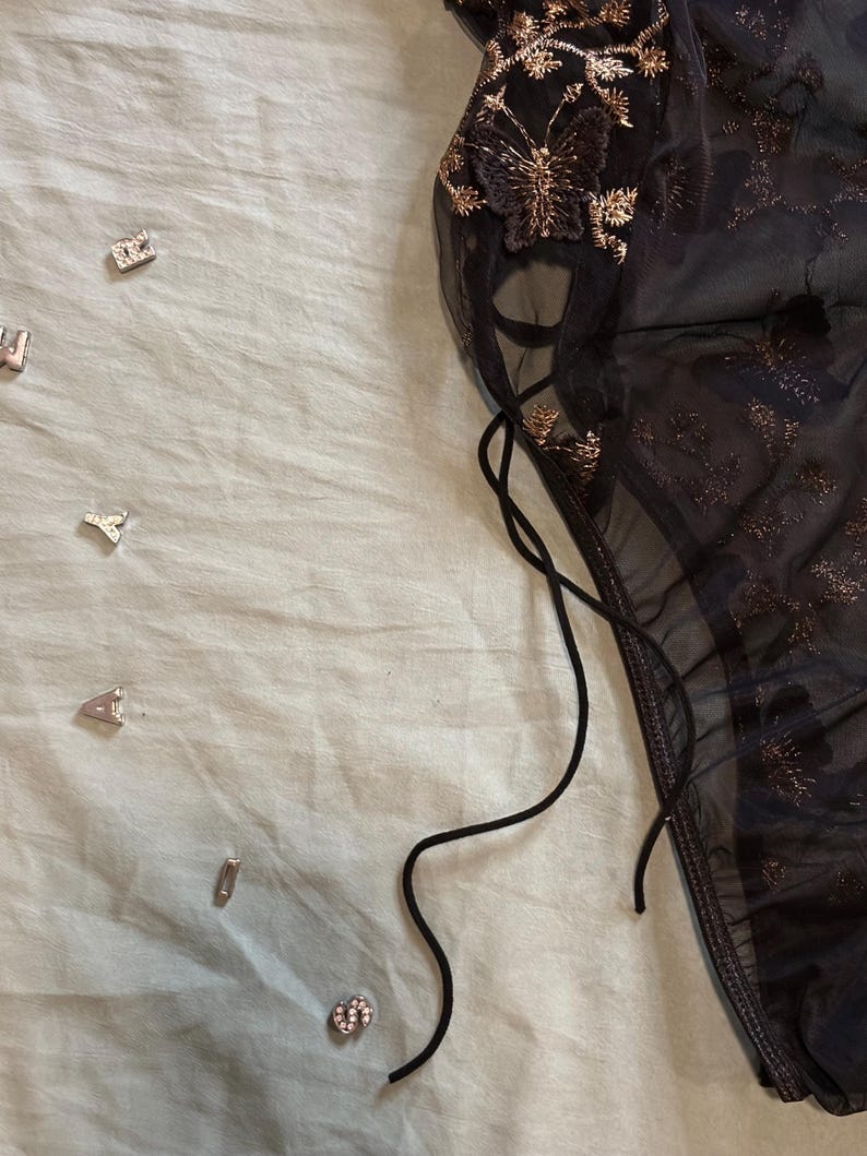 May include: A sheer black lingerie piece with gold butterfly and floral embroidery. The garment features delicate lace detailing and a drawstring. Scattered around are small silver-colored decorative charms.