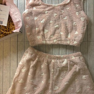 K&ouml;nnte beinhalten: Ein rosa, flauschiges Pyjama-Set mit wei&szlig;en Herzmustern. Das Set besteht aus einem Crop-Top und Shorts, beide aus einem weichen, strukturierten Material. Der Pyjama wird auf einer neutralen Oberfl&auml;che pr&auml;sentiert.