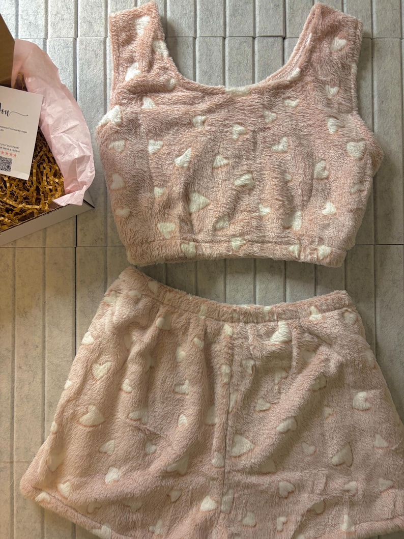 K&ouml;nnte beinhalten: Ein rosa, flauschiges Pyjama-Set mit wei&szlig;en Herzmustern. Das Set besteht aus einem Crop-Top und Shorts, beide aus einem weichen, strukturierten Material. Der Pyjama wird auf einer neutralen Oberfl&auml;che pr&auml;sentiert.