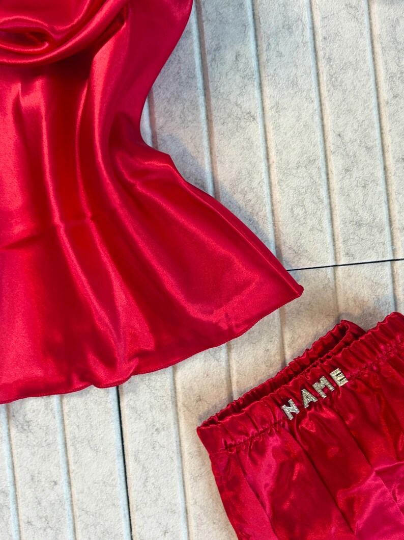 May include: A red satin pajama set. The top is a draped style, and the shorts have an elastic waistband with the word "NAME" spelled out in silver rhinestones. The fabric has a shiny, smooth texture.