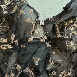 May include: Close-up of a black sheer garment with gold embroidered butterfly and floral designs. The garment has a V-neckline and a small metal detail with the letter "N". The fabric is delicate and detailed.
