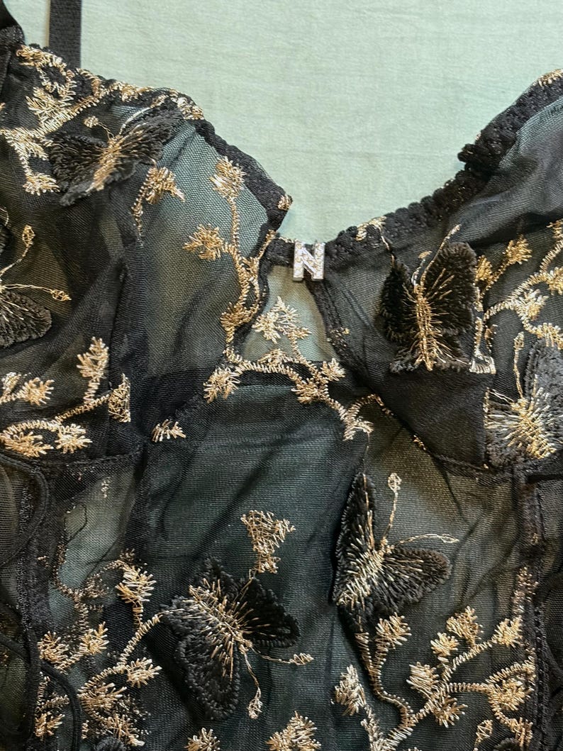 May include: Close-up of a black sheer garment with gold embroidered butterfly and floral designs. The garment has a V-neckline and a small metal detail with the letter "N". The fabric is delicate and detailed.