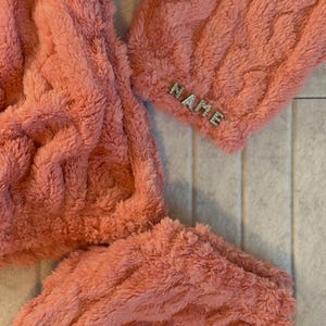May include: Close-up of a coral-colored, plush, cable-knit fabric. The word "NAME" is spelled out in small, silver-colored embellishments on the cuff of the garment. The fabric appears soft and cozy.
