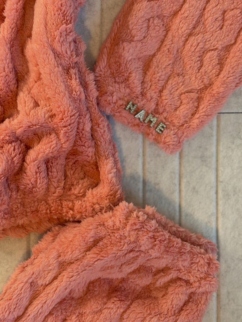 May include: Close-up of a coral-colored, plush, cable-knit fabric. The word "NAME" is spelled out in small, silver-colored embellishments on the cuff of the garment. The fabric appears soft and cozy.