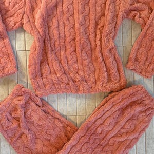 May include: A coral-colored, plush pajama set featuring a cable knit design. The set includes a long-sleeved top and matching pants. The fabric appears soft and comfortable, ideal for lounging or sleeping. The set is laid out on a tiled surface.
