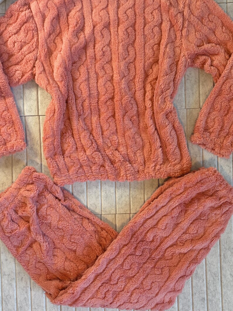 May include: A coral-colored, plush pajama set featuring a cable knit design. The set includes a long-sleeved top and matching pants. The fabric appears soft and comfortable, ideal for lounging or sleeping. The set is laid out on a tiled surface.