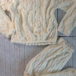 May include: Cream-colored, plush loungewear set. The set includes a long-sleeved top and matching pants. The fabric has a textured, cable-knit appearance. The top has a gathered waist and the pants are folded. The set is displayed on a light gray tiled surface.
