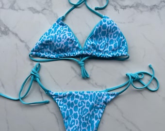 May include: A two-piece bikini set featuring a blue and white leopard print. The triangle top and the bottom have tie closures. The bikini is displayed on a white marble surface.