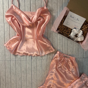 Op de afbeelding: Een perzikkleurige satijnen pyjamaset, inclusief een camisole top met spaghettibandjes en een watervalhalslijn, en een bijpassende pyjamabroek. Een geschenkdoos met een kaart en scrunchie is ook inbegrepen.