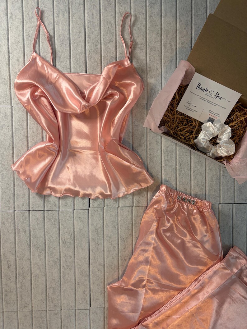 Op de afbeelding: Een perzikkleurige satijnen pyjamaset, inclusief een camisole top met spaghettibandjes en een watervalhalslijn, en een bijpassende pyjamabroek. Een geschenkdoos met een kaart en scrunchie is ook inbegrepen.