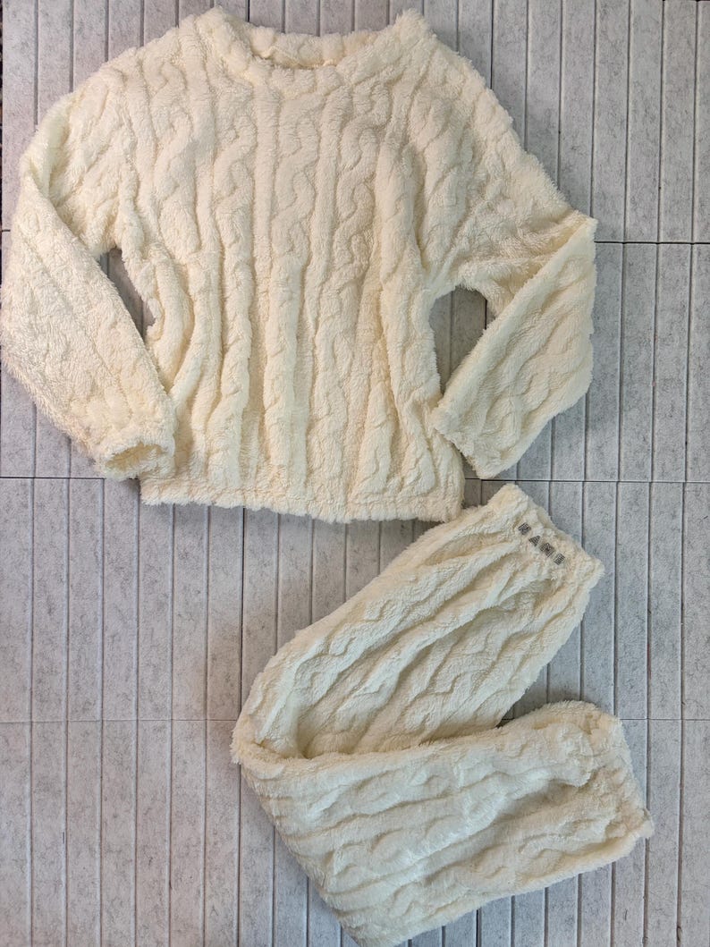 May include: Cream-colored, cable-knit pajama set. The set includes a long-sleeved top and matching pants. The fabric appears soft and plush, ideal for lounging. The top has a crew neck, and the pants have a drawstring waist.