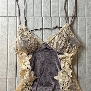 May include: A camisole featuring a dark gray velvet bodice with cream-colored lace overlays on the cups and sides. The garment includes thin adjustable shoulder straps, a small decorative accent at the center neckline, and floral lace appliques.
