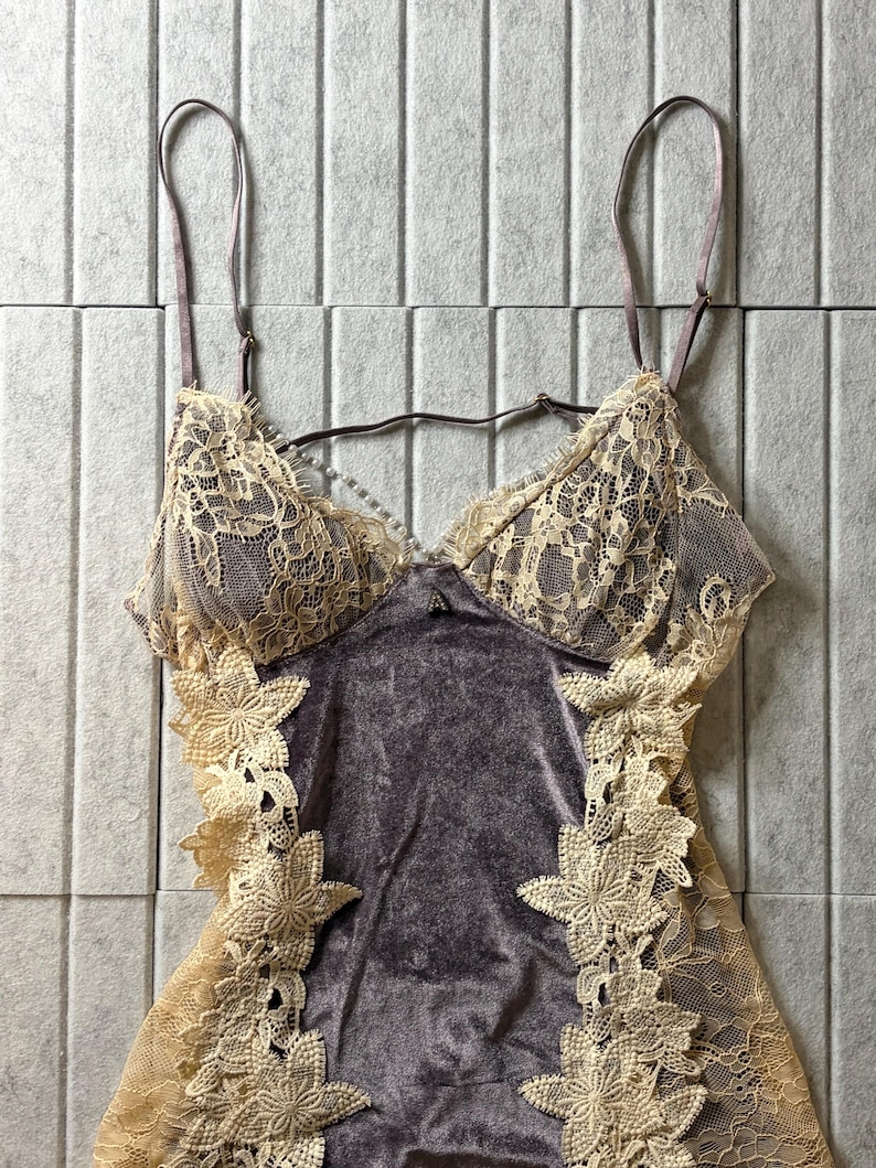 May include: A camisole featuring a dark gray velvet bodice with cream-colored lace overlays on the cups and sides. The garment includes thin adjustable shoulder straps, a small decorative accent at the center neckline, and floral lace appliques.