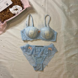 May include: A light blue lingerie set with lace detailing and small peach-colored bows. The set includes a bra and matching panties. A white scrunchie and a gift box with a card are also visible.
