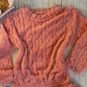 May include: A coral-colored, plush sweater and matching bottoms set. The sweater features a cable knit design and long sleeves. The word "NAME" is visible on the cuff of the sleeve. The set appears soft and comfortable.