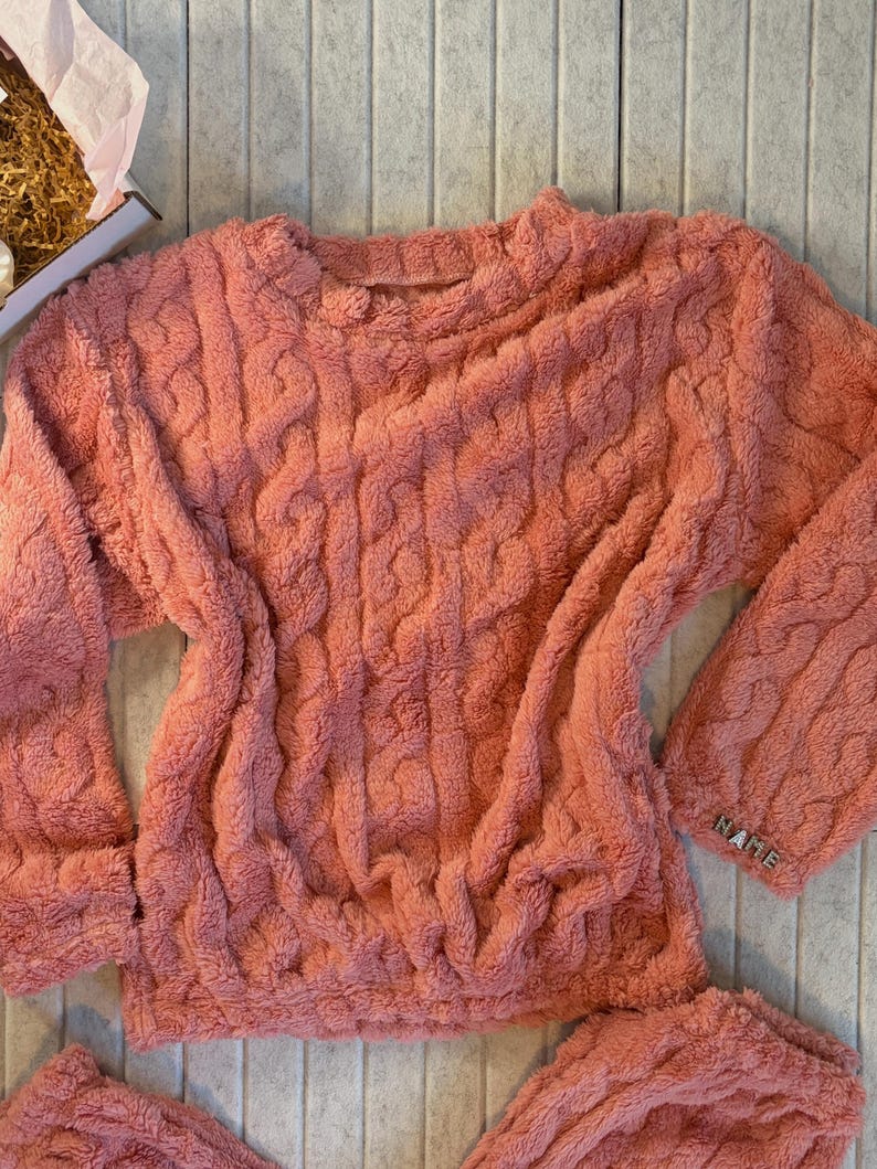 May include: A coral-colored, plush sweater and matching bottoms set. The sweater features a cable knit design and long sleeves. The word "NAME" is visible on the cuff of the sleeve. The set appears soft and comfortable.