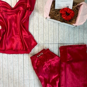 May include: A red satin pajama set with a camisole top and pants, along with a matching scrunchie and a gift box. The pants have the word "NAME" embroidered on the waistband. The gift box contains a thank you card.