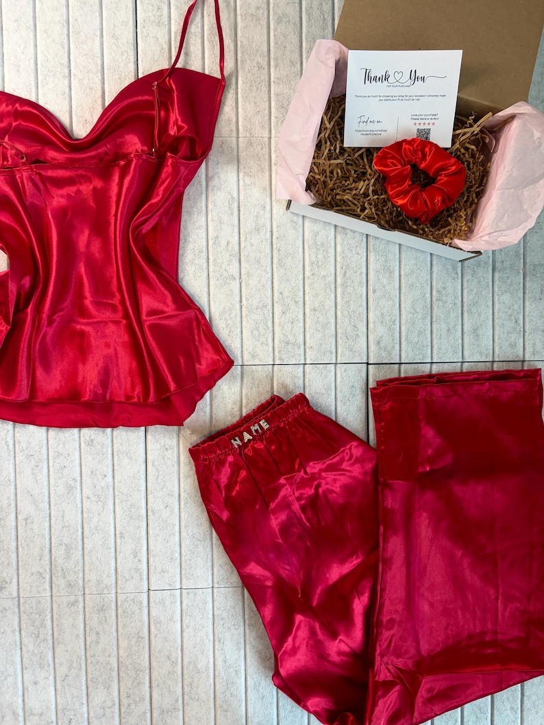May include: A red satin pajama set with a camisole top and pants, along with a matching scrunchie and a gift box. The pants have the word "NAME" embroidered on the waistband. The gift box contains a thank you card.