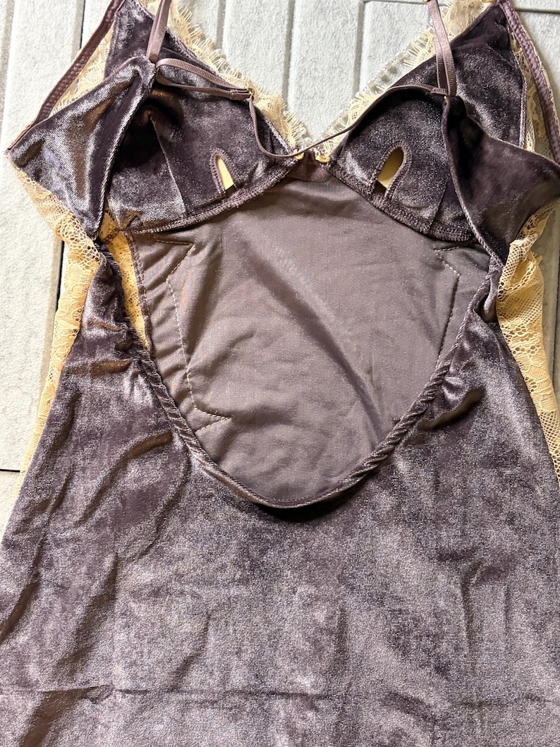 May include: A dark gray velvet camisole featuring delicate beige lace trim along the neckline and sides. The garment includes thin adjustable shoulder straps and a central panel with a smooth fabric finish, showcasing a combination of soft textures.