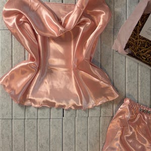 Op de afbeelding: Een lichtroze satijnen pyjamaset, bestaande uit een camisole top met een watervalhalslijn en bijpassende shorts. De set wordt gepresenteerd op een gestructureerd, lichtgrijs oppervlak, met een geschenkdoos op de achtergrond.