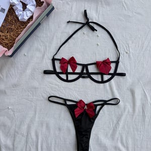May include: A black lingerie set consisting of a strappy underwire bra and a thong. The bra features two red satin bows on the cups and thin black straps. The thong includes a matching red satin bow at the center with a small letter A charm.