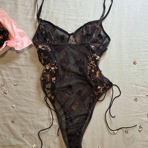 May include: Black lingerie bodysuit with gold floral embroidery. The sheer fabric features adjustable straps and side ties. A pink tissue-lined box is visible in the upper left corner, with scattered letter charms on a light green surface.