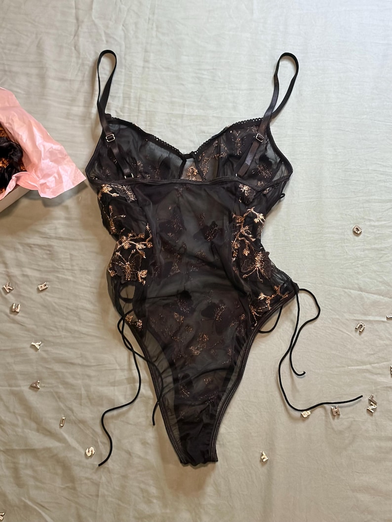 May include: Black lingerie bodysuit with gold floral embroidery. The sheer fabric features adjustable straps and side ties. A pink tissue-lined box is visible in the upper left corner, with scattered letter charms on a light green surface.