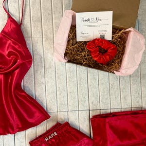 May include: A red satin pajama set with a matching camisole and shorts, along with a red scrunchie and a gift box. The shorts have the word "NAME" embroidered on the waistband. A "Thank You" card is inside the box.