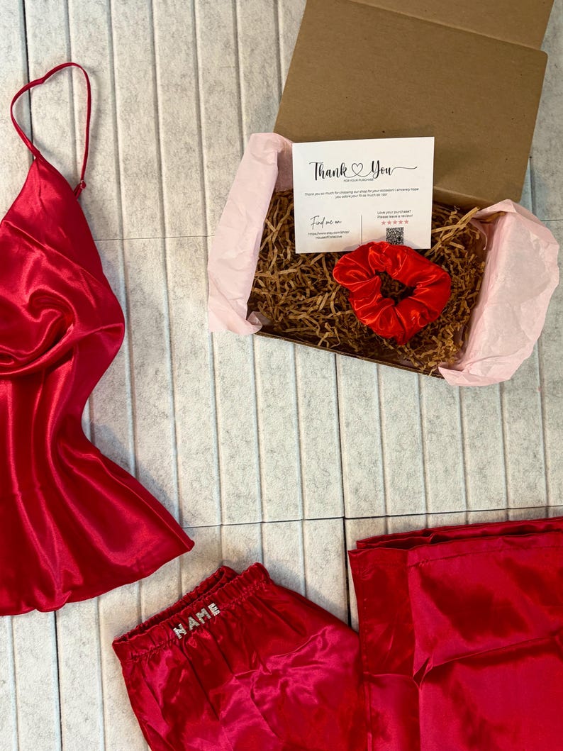 May include: A red satin pajama set with a matching camisole and shorts, along with a red scrunchie and a gift box. The shorts have the word "NAME" embroidered on the waistband. A "Thank You" card is inside the box.