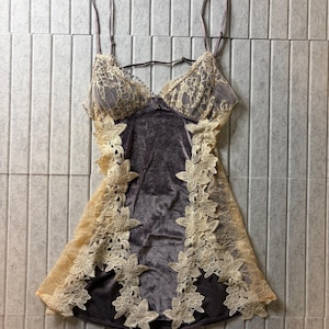 May include: A dark gray velvet chemise featuring beige floral lace trim along the cups and sides. The garment has thin adjustable shoulder straps, a V-neckline, and a fitted silhouette with intricate lace flower appliqu&eacute;s bordering the velvet.
