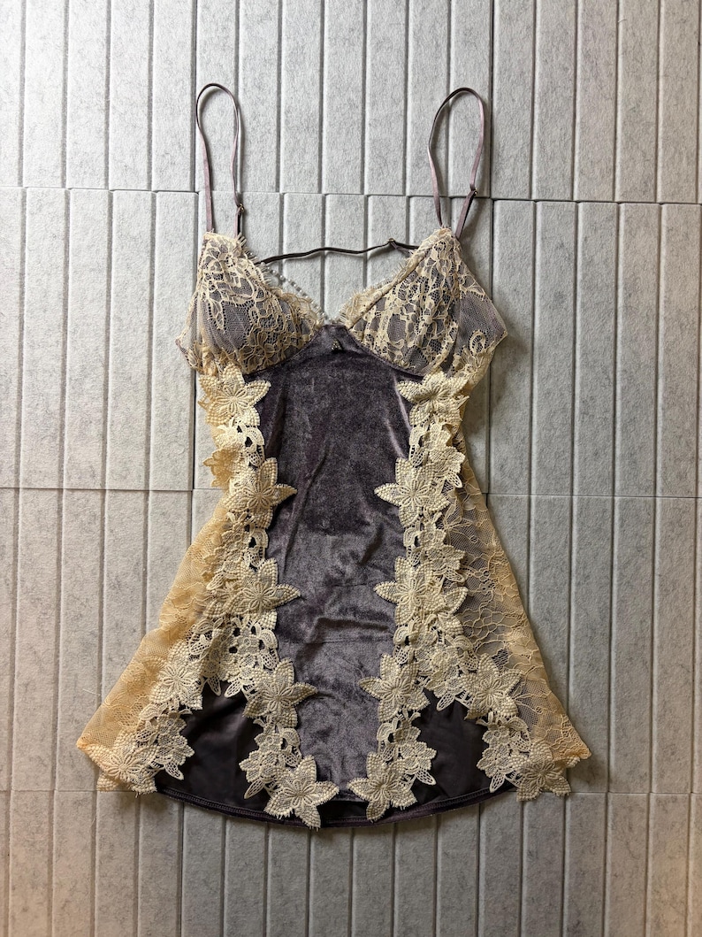 May include: A dark gray velvet chemise featuring beige floral lace trim along the cups and sides. The garment has thin adjustable shoulder straps, a V-neckline, and a fitted silhouette with intricate lace flower appliqu&eacute;s bordering the velvet.